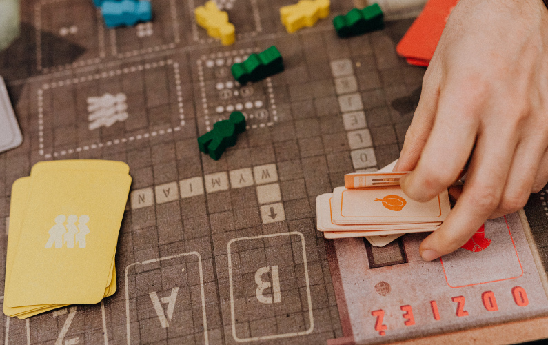 STEM Mondays.png Image Breakdown Long Description: The image displays a section of a board game laid out on a tabletop. In the foreground, a person’s hand is arranging a stack of cream-colored cards with an orange vegetable silhouette on the top card. To the left, there is a pile of yellow cards featuring a simplistic human icon in white. The board has a grid-like structure with different sections marked with text and graphics. Several small, colored wooden tokens are scattered across the board, including green, yellow, and blue figures. The background has a mix of dark and light brown tones, and various text elements are printed on the board. Alt-text: A hand arranges cards on a board game with colored tokens.